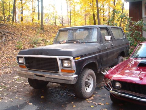 1978 Ford Bronco - Flat-black full-size Ford Bronco with a hardtop parked on a leaf-strewn driveway in autumn woods, shot from the driver's front three-quarte…