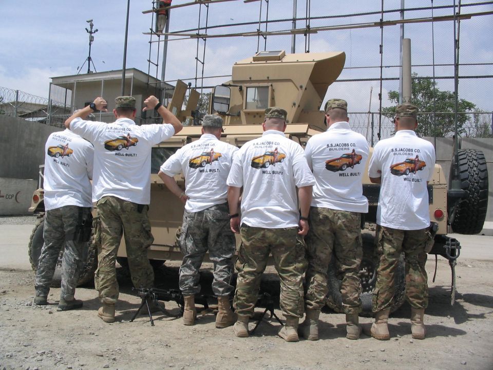 Soldiers in front of a tan military vehicle, some wearing shirts with a car graphic