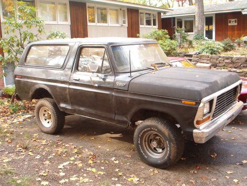 1978 Ford Bronco - A flat-black full-size Ford Bronco Custom sits in a leaf-covered driveway beside a wood-sided house, shown from the front three-quarter wit…