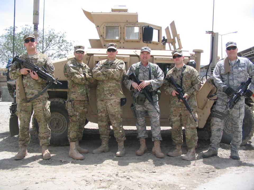Six soldiers in camouflage stand in front of a tan armored vehicle