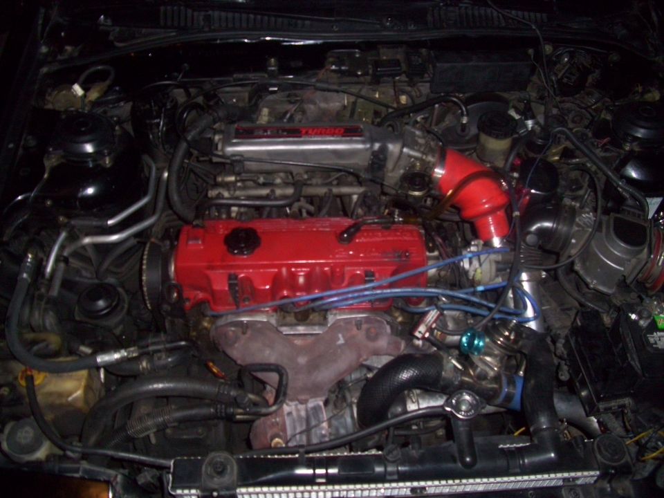 Engine bay with a red valve cover, turbocharger, and red intake piping. Text on the bottom reads 'SUPERMOTORS'