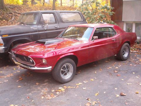 1978 Ford Bronco - Three-quarter front view of a candy-red 1969 Ford Mustang notchback coupe with a black hood scoop stripe and polished five-spoke mag wheels…