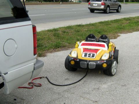 1999 Ford Ranger - A small yellow toy car is being towed by a larger vehicle on a paved surface next to a grassy area.