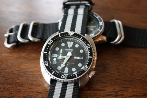 1986 Ford Bronco - A close-up view of a Seiko automatic watch with a black and gray striped NATO strap, resting on a wooden surface.