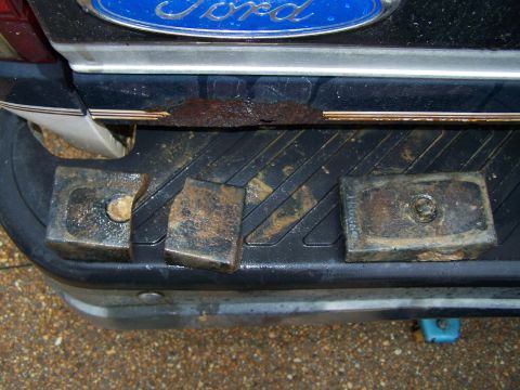 1992 Ford Bronco - Four metal lift blocks are placed on the textured rear bumper of a Ford vehicle, which shows signs of rust.