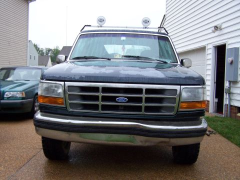 1992 Ford Bronco - A dark blue 1992 Ford Bronco is parked in a driveway next to a house, with a green Volvo station wagon visible on the left.