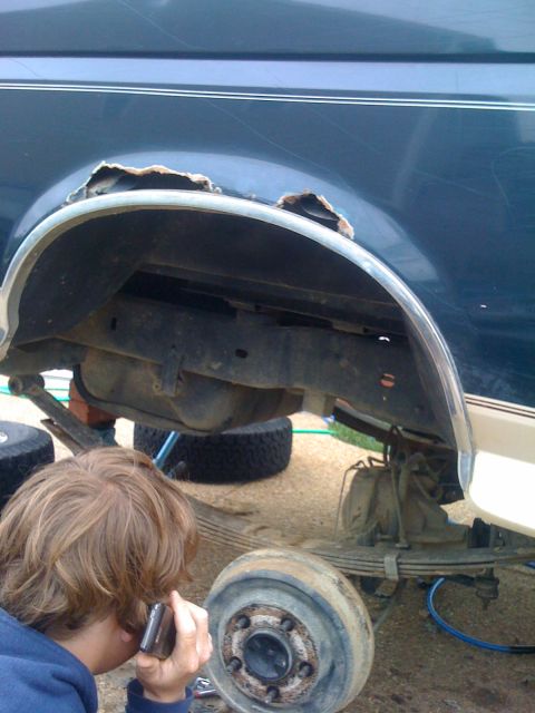 1992 Ford Bronco - The rear wheel well of a dark blue SUV shows significant rust damage. Suspension parts are visible below.