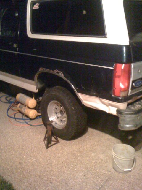 1992 Ford Bronco - The rear of a dark blue and white Ford Bronco is shown with a jack supporting the rear wheel and air compressors on the ground.