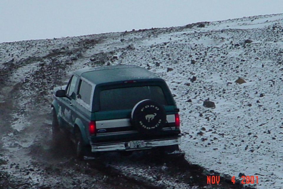 Green Ford Bronco climbing snowy rocky trail, rear view with spare tire cover.