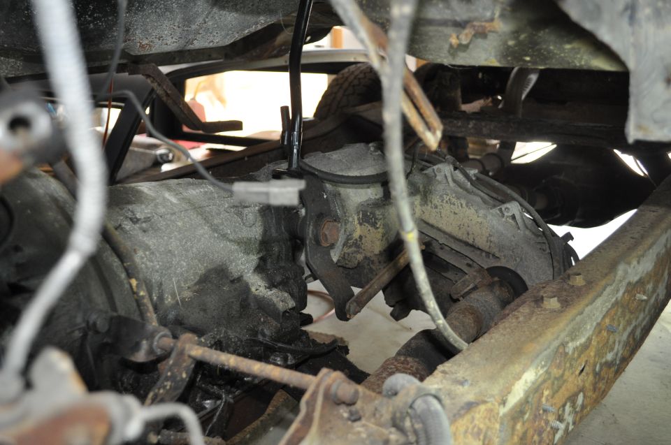 Undercarriage view of a vehicle showing transmission, driveshaft, and suspension parts. Some rust is visible on the frame
