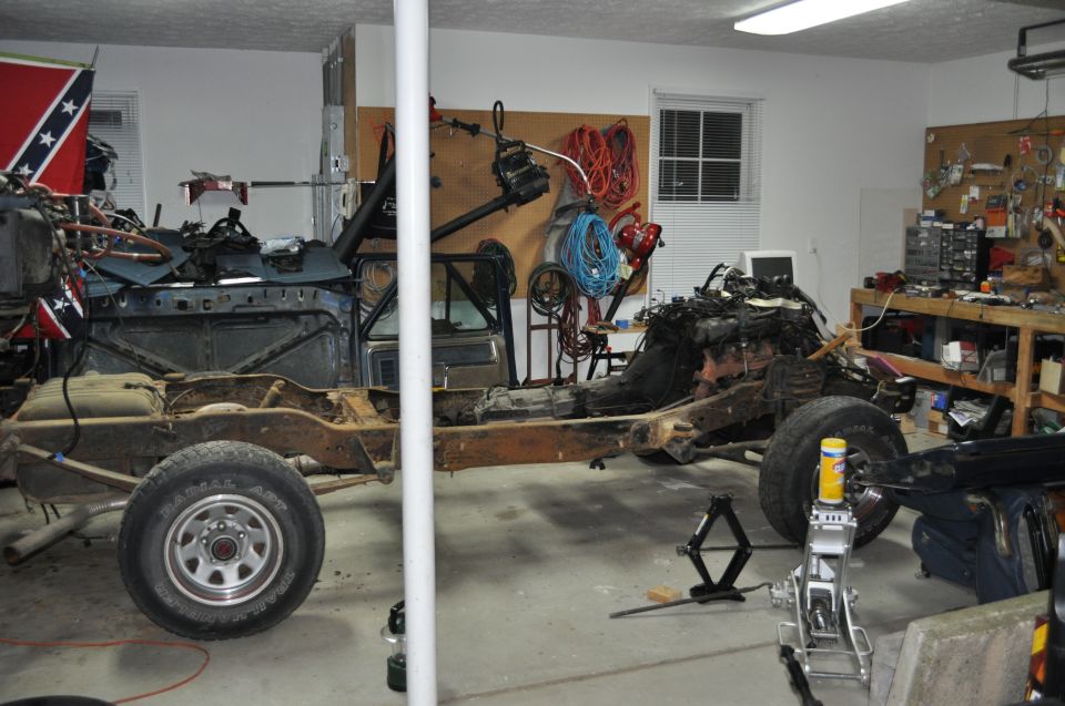vehicle frame with engine and suspension parts, in a garage with tools and a workbench