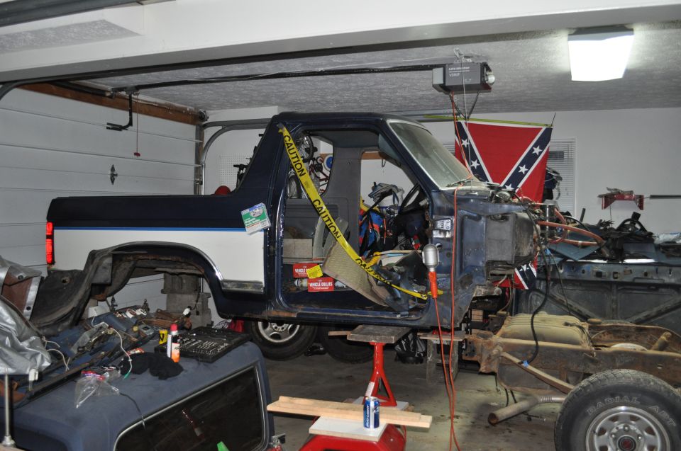 Disassembled blue and white Ford Bronco in garage, front end removed, on jack stands