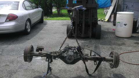 1987 Ford Ranger - A detached rear axle assembly is hoisted by chains, with a silver sedan and a white barrel in the background.