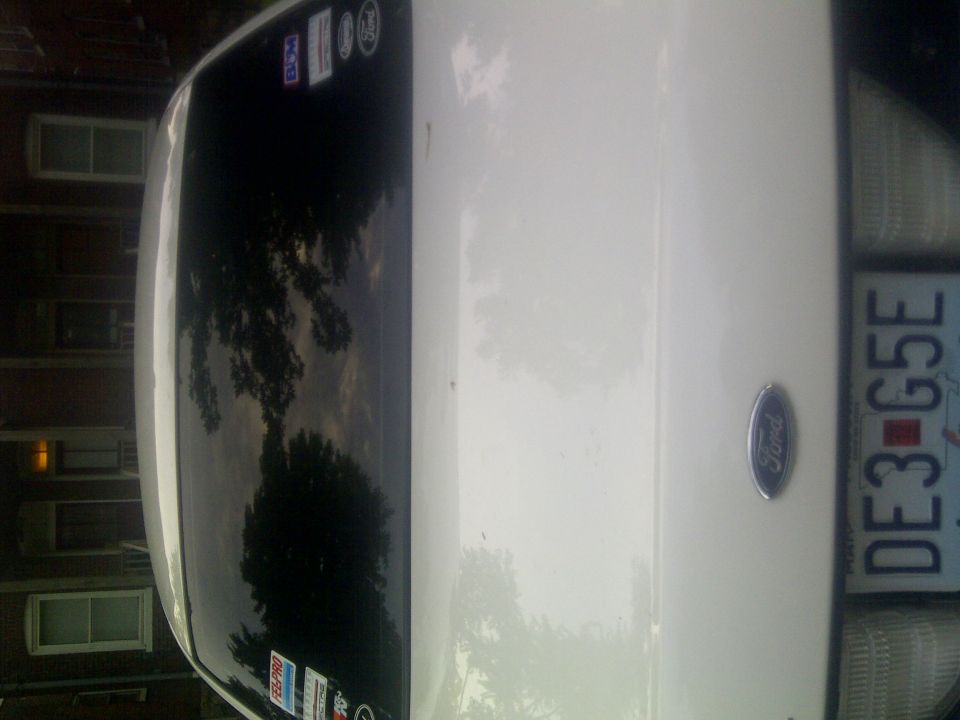 Rear of a white 2000 Ford Crown Victoria, showing the trunk lid, taillight, license plate DE3-G5E, and stickers on the rear window