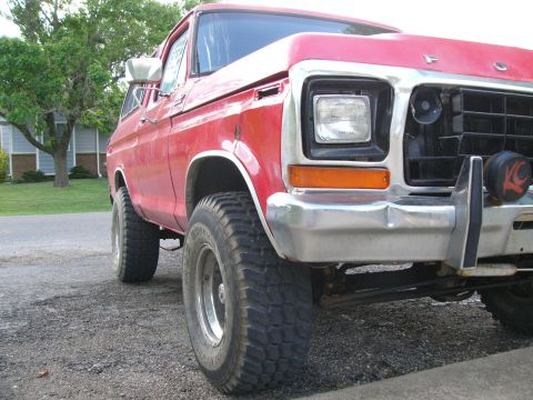1979 Ford Bronco - A red Ford Bronco sits parked on a gravel surface, with a house and trees in the background.