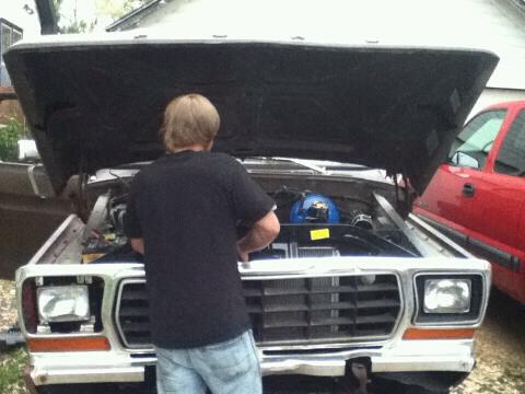 1979 Ford Bronco - A man works on the engine of a 1979 Ford Bronco, with a red truck parked nearby.