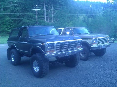 1979 Ford Bronco - Two Ford Broncos are parked outdoors, with a wooded area in the background. The black Bronco is in the foreground.