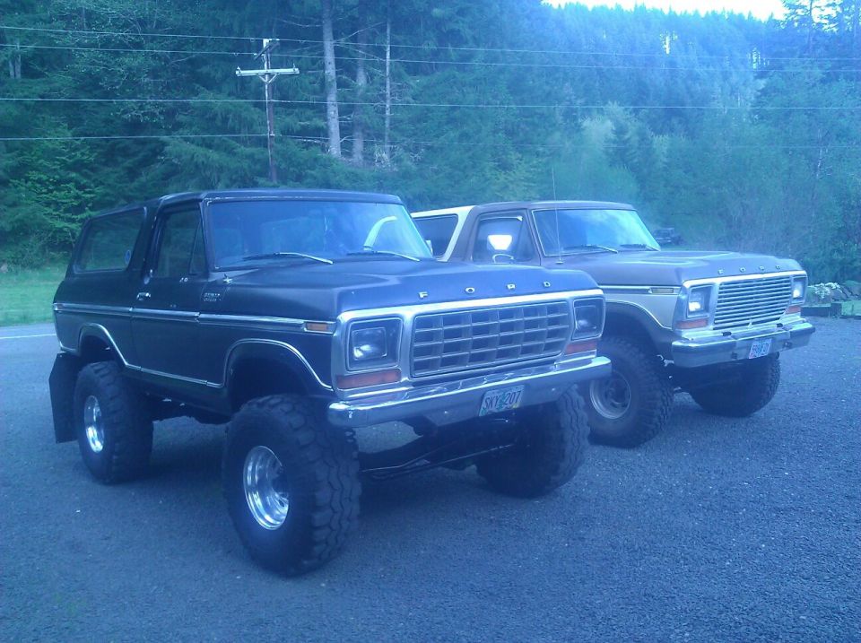 Two lifted Ford Broncos parked side-by-side, one black and one brown. The black Bronco is in the foreground