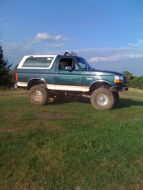 1992 Ford Bronco - Modified 1992 Ford Bronco with off-road tires and roof-mounted lights sits on a grassy knoll under a blue sky.