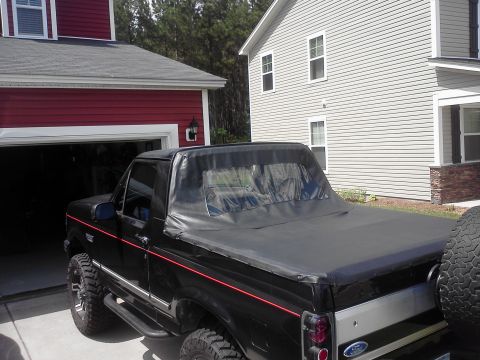 1992 Ford Bronco - This black Ford Bronco features a soft top and is parked in a driveway next to a house.