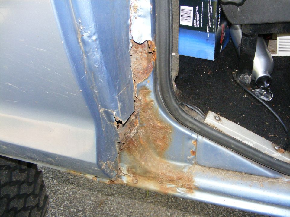rusted out door frame of a blue vehicle, showing significant rust damage around the hinges and lower panel. Tire tread visible