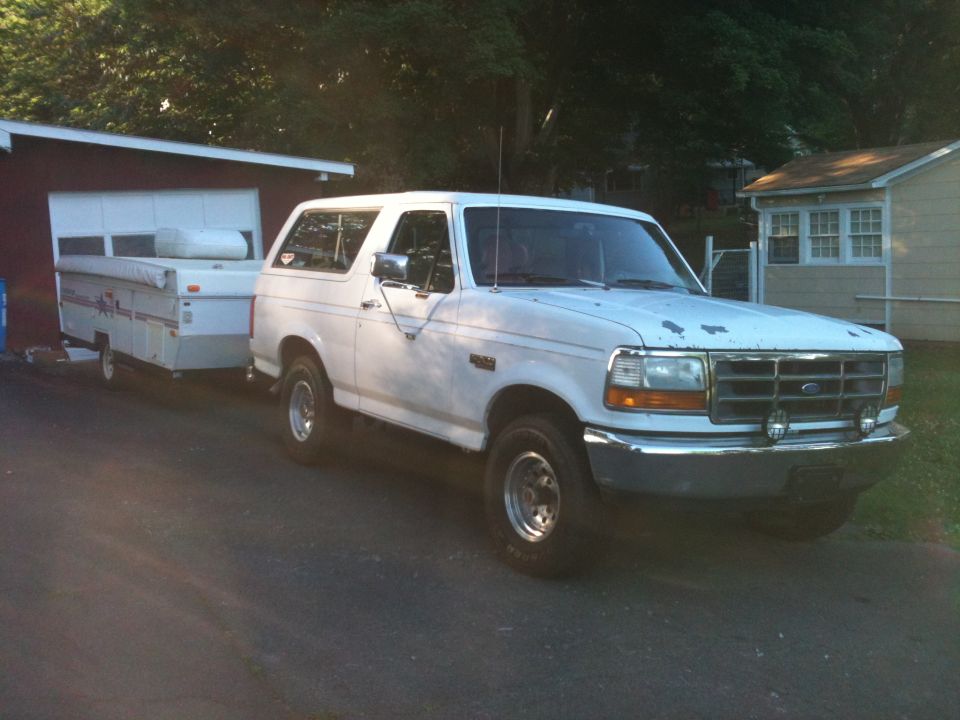 White 1992 Ford Bronco with rust spots on the hood, towing a white pop-up camper