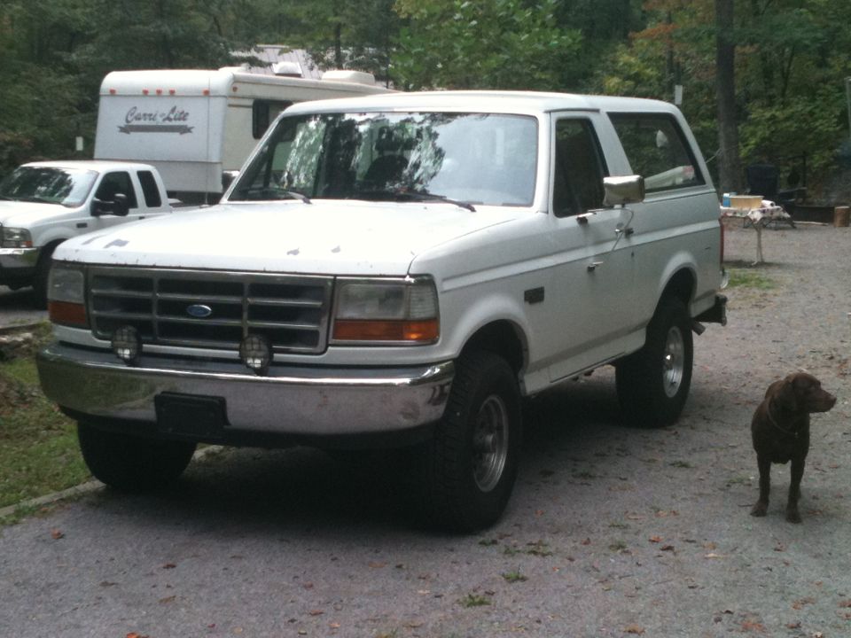1992 Ford Bronco parked next to a white pickup truck and a camper trailer, with a brown dog standing nearby