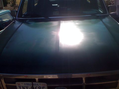 1992 Ford Bronco - A dark green SUV's hood and windshield are visible, with a bright sun glare obscuring details.