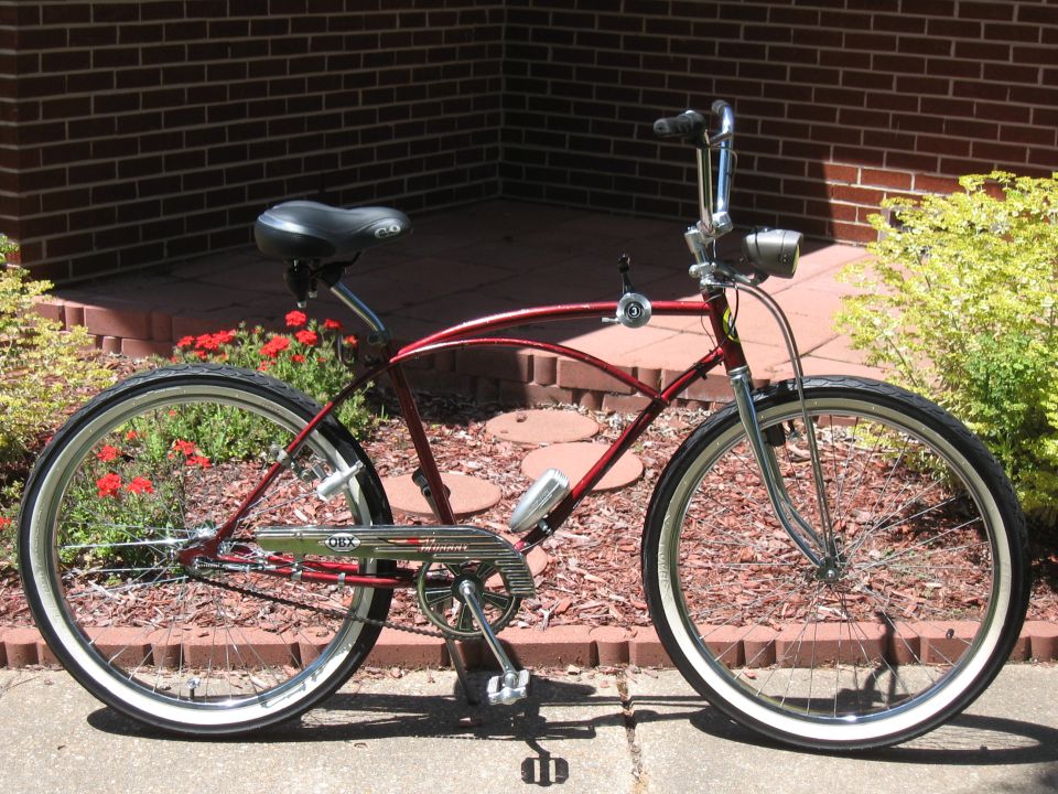 Red cruiser bicycle with chrome handlebars, fenders, and chain guard. Has whitewall tires and a headlight