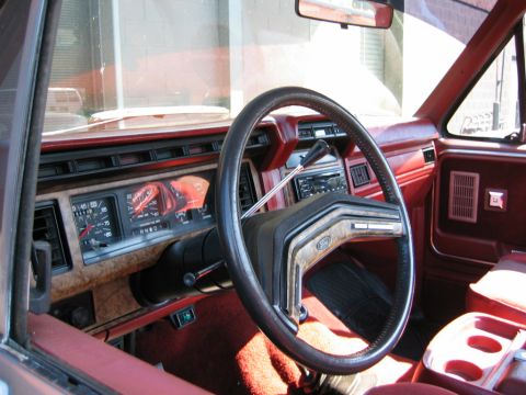 1986 Ford Bronco - Red interior of a Ford truck shot from the open driver's door, showing the black four-spoke steering wheel with Ford emblem, woodgrain dash…
