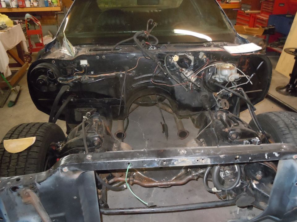 Disassembled car engine bay, dark blue exterior, front view, showing engine mounts and exhaust pipes