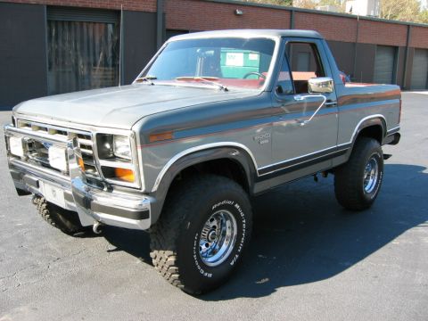 1986 Ford Bronco - Driver-side three-quarter front view of a gray 1980s Ford Bronco with its rear hardtop removed, showing a red interior. It wears a chrome g…