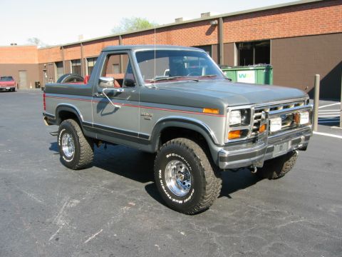 1986 Ford Bronco - Passenger-side three-quarter front view of a gray 1980s Ford Bronco with the rear top removed, red pinstriping, a chrome brush guard with t…