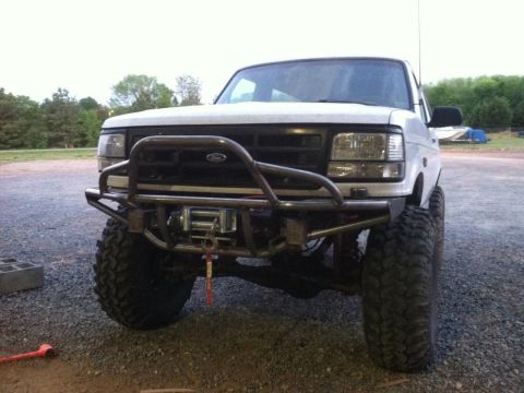 1996 Ford Bronco - Front three-quarter view of a lifted white Ford Bronco on gravel, wearing a bare-steel tube prerunner bumper with hoop and a winch mounted…