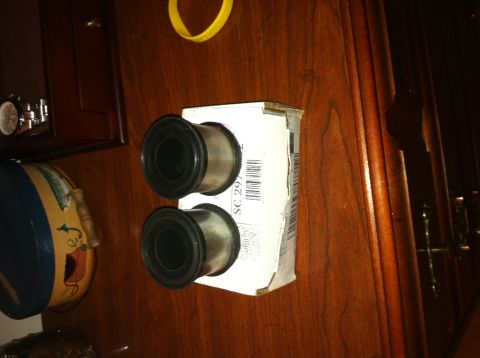 1996 Ford Bronco - Two new suspension control-arm bushings with steel outer sleeves and black rubber flanges standing on their white parts box, which is label…