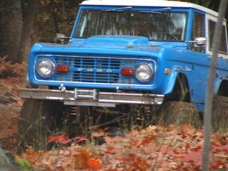 1989 Ford Bronco - A blue early Ford Bronco with a white roof climbs a leaf-covered bank in the woods, shot head-on with the front end lifted and the tires fl…