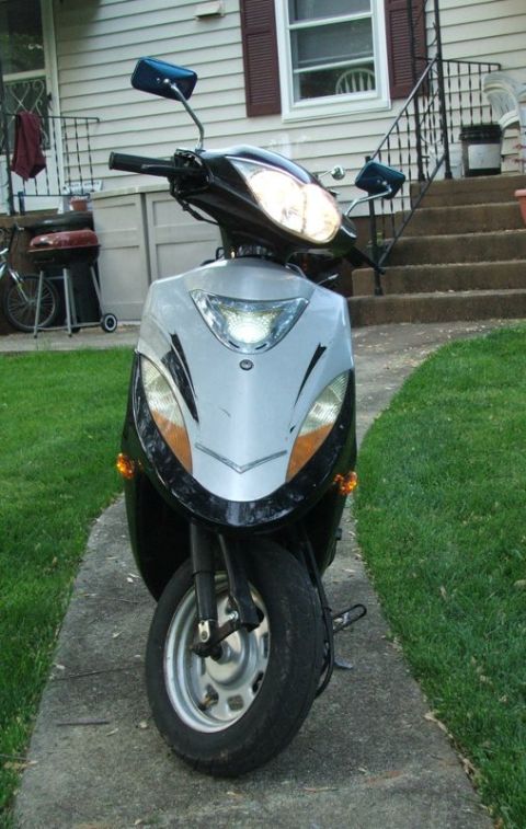 2008 Yamaha Zuma - A scooter is parked on a walkway in front of a residential home, with a grill visible to the left.
