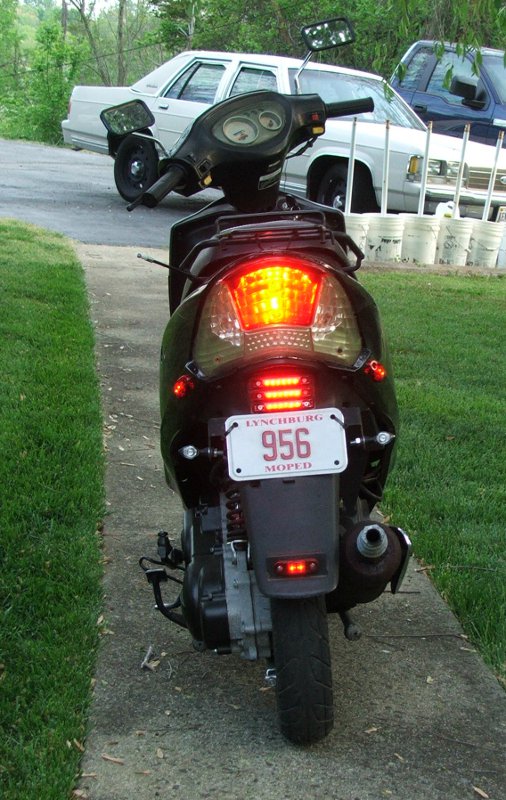 Black scooter rear view, license plate 956 MOPED, with two cars in background