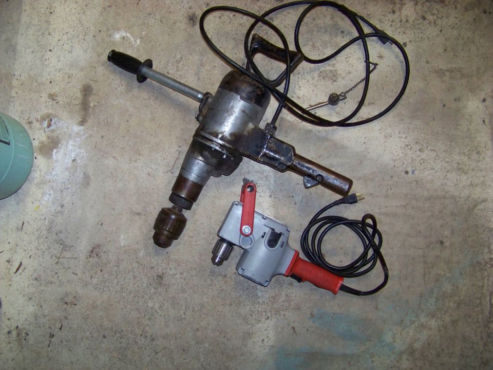 Two electric drills, one large and old, the other smaller and red, sit on a concrete floor