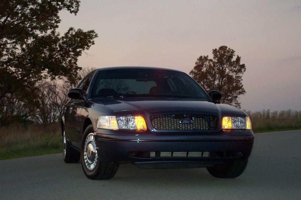 Dark maroon Ford Crown Victoria sedan front three-quarter at dusk, headlights on, on a country road
