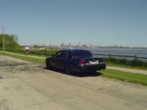 1999 Ford Crown Victoria - A dark sedan is parked near a waterfront with a city skyline visible across the water.