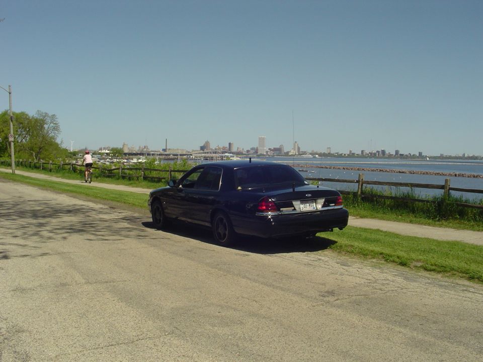 Dark sedan parked on a road next to a body of water with a city skyline in the distance