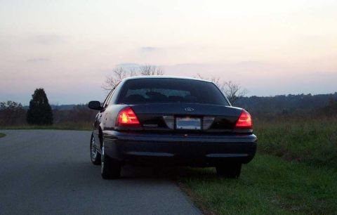 1998 Ford Crown Victoria - Rear view of a dark-colored Ford Crown Victoria full-size sedan parked on a paved road at dusk, with its taillights illuminated and a rural…
