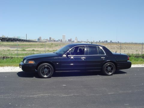1999 Ford Crown Victoria - A black sedan is parked on a road with a city skyline visible in the distance.