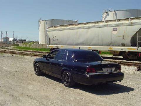 1999 Ford Crown Victoria - A black sedan is parked on a dirt road next to a train car under a clear blue sky.