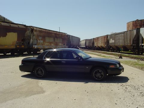 1999 Ford Crown Victoria - A black 1999 Ford Crown Victoria is seen from the side, parked next to a line of train cars under a clear blue sky.
