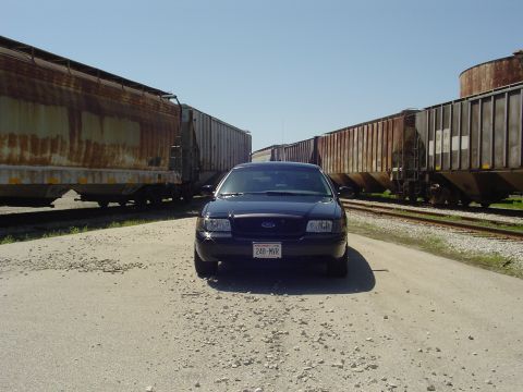 1999 Ford Crown Victoria - Dark sedan parked on gravel road between two long train cars. License plate reads 248-MVR.