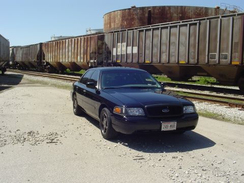 1999 Ford Crown Victoria - A black sedan is parked next to a long train on a sunny day.