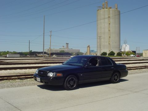 1999 Ford Crown Victoria - Black sedan parked on concrete near railroad tracks with industrial buildings in background.