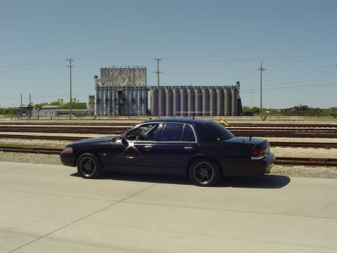 1999 Ford Crown Victoria - A black sedan is parked next to train tracks with large industrial silos in the distance.